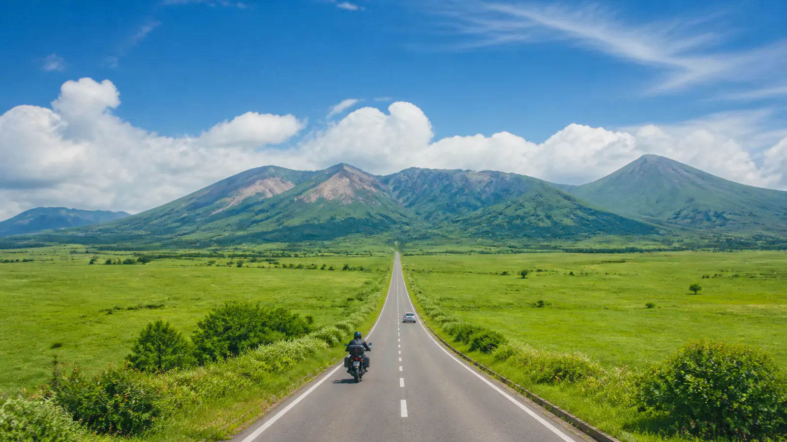 くじゅうの山並みへ伸びる草原の道を車とバイクが走るイメージ